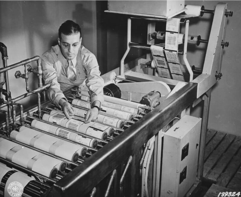 A man is operating a vintage printing machine, carefully aligning several rolls of paper. The machine is large, with multiple rollers and technical components. The setting appears to be a workplace or industrial environment.